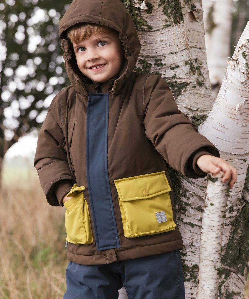 Dreng i varmt overtøj med hætte står ved et træ udendørs.