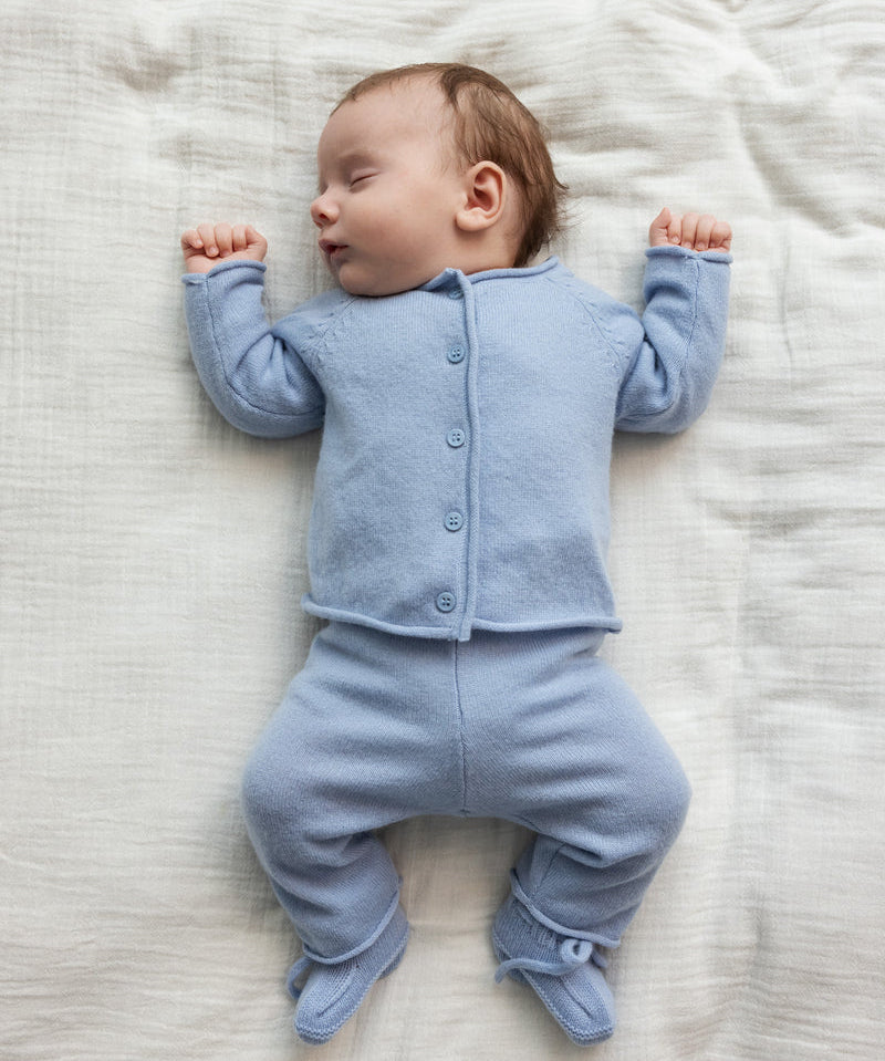 Sovende baby iført lyseblåt striksæt med cardigan, bukser og futter liggende på blødt tæppe.