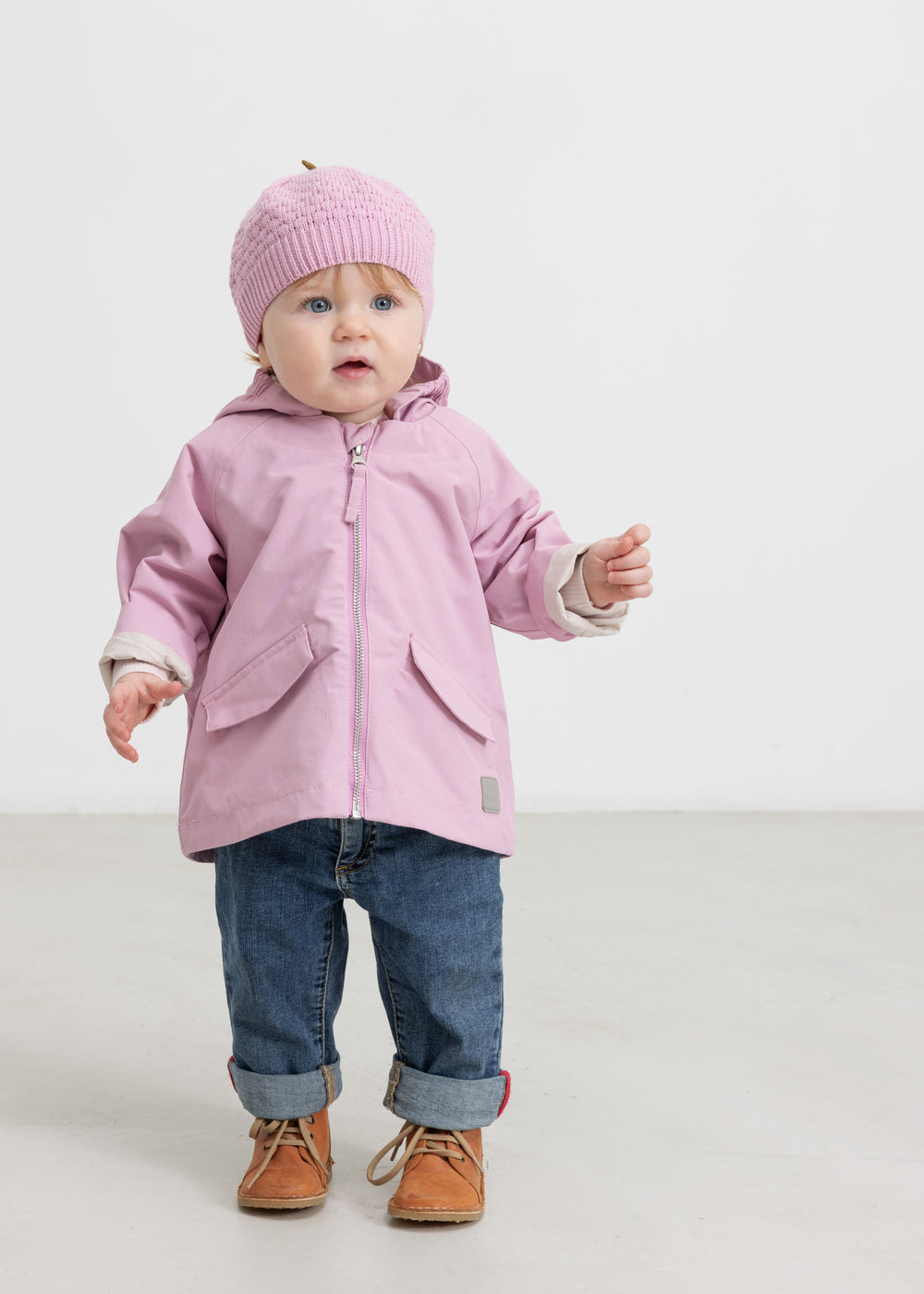 Baby iført lyserød overgangsjakke, rosa hue og jeans
