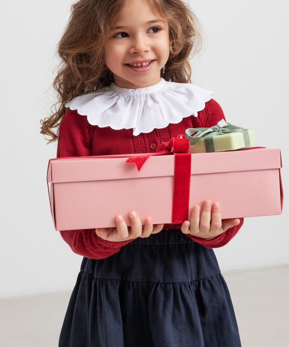 Pige iført rød juletrøje med hulmønster og hvid krave og blå nederdel, holder julegaver