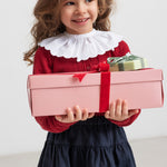 Pige iført rød juletrøje med hulmønster og hvid krave og blå nederdel, holder julegaver