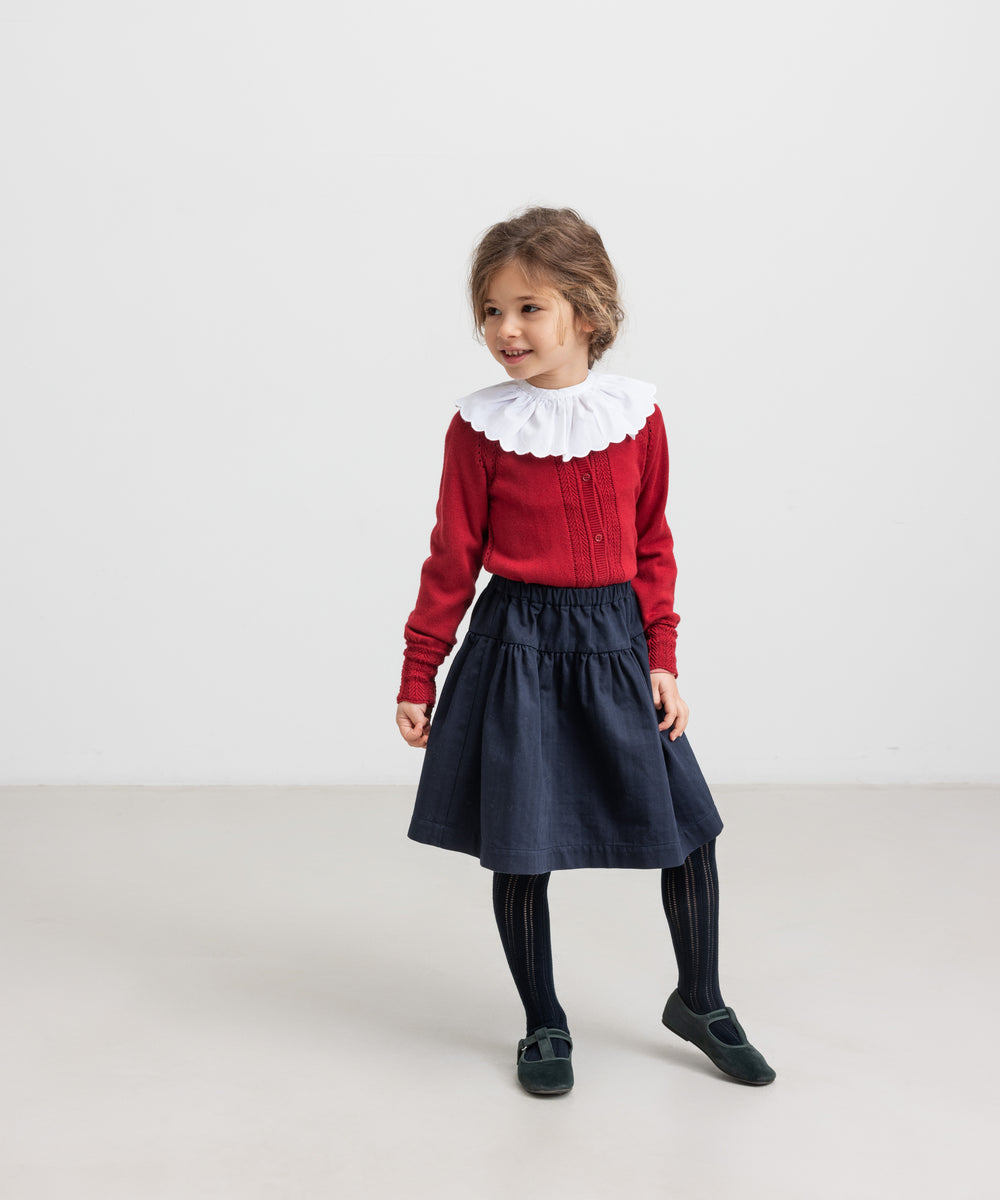 Pige iført mørkeblåt nederdel, rød bluse med hvid krave og sorte strømpebukser til børn