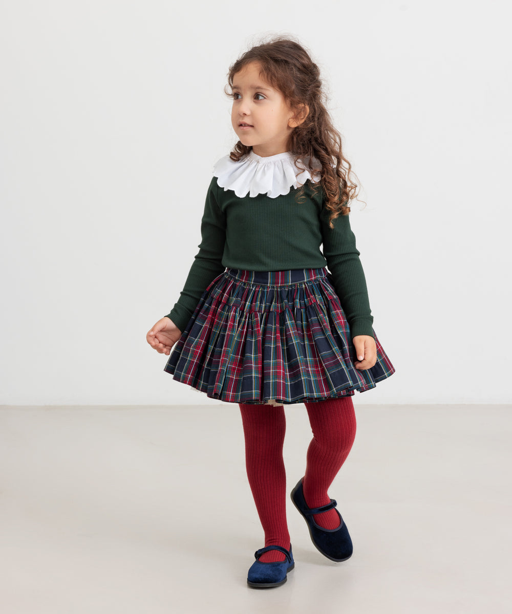 Pige iført juletøj - mørkeblåt og rødt ternet nederdel, mørkegrøn bluse og røde strømpebukser til børn