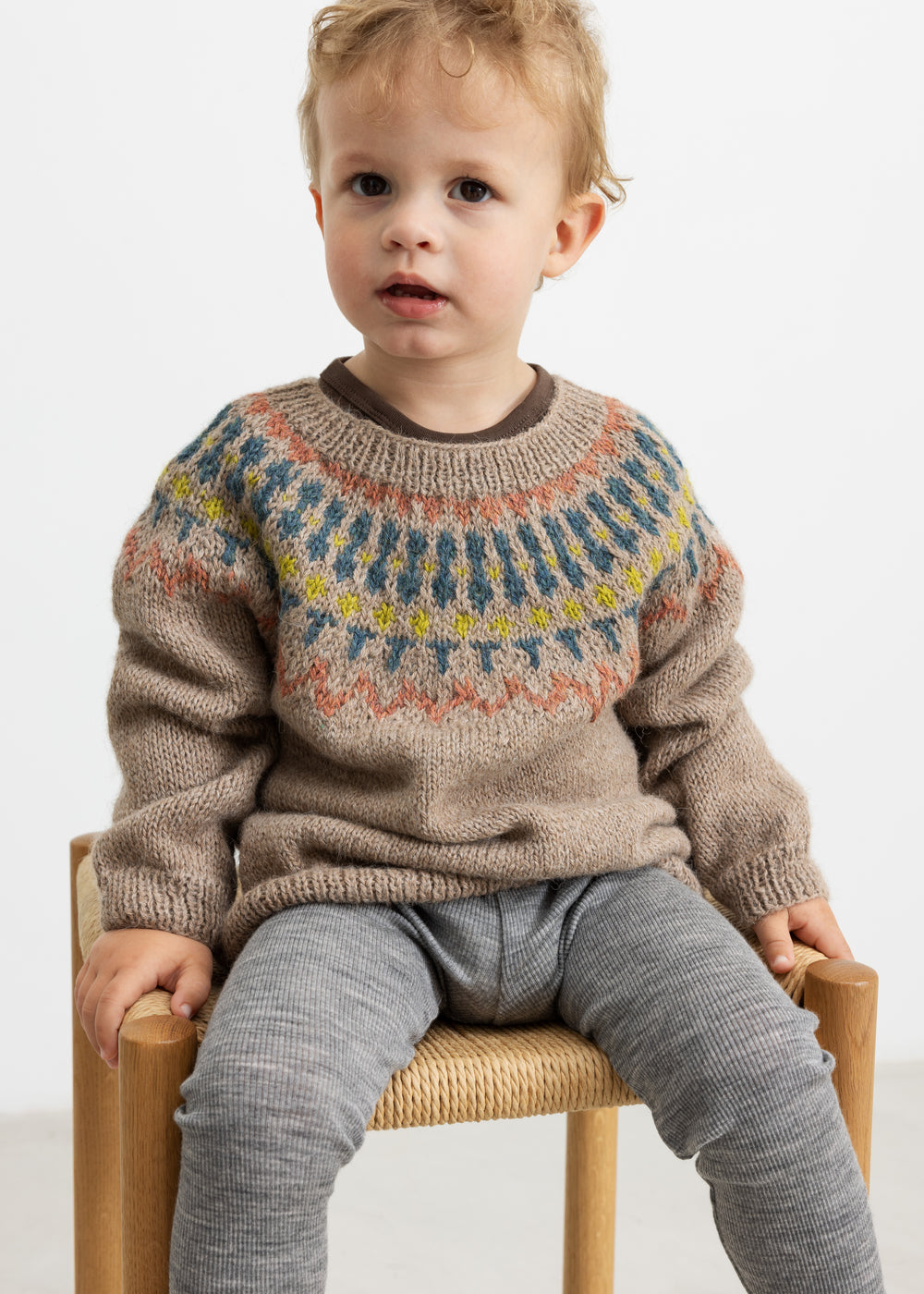 Dreng iført beige uldstrikbluse med mønster sidder på stol
