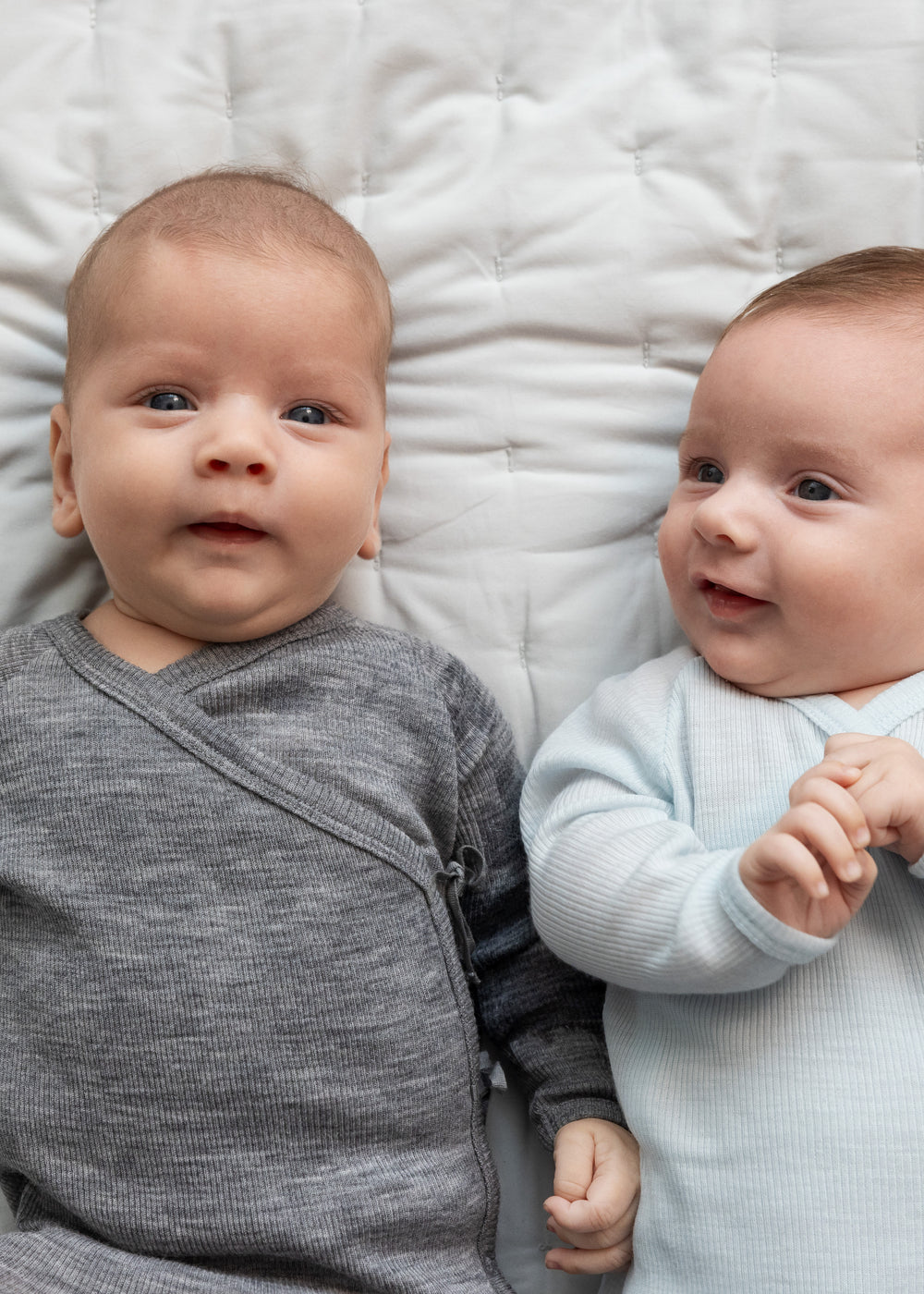 To babyer ligger på seng iført heldragter i merinould, en grå og en lyseblå