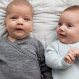 To babyer ligger på seng iført heldragter i merinould, en grå og en lyseblå