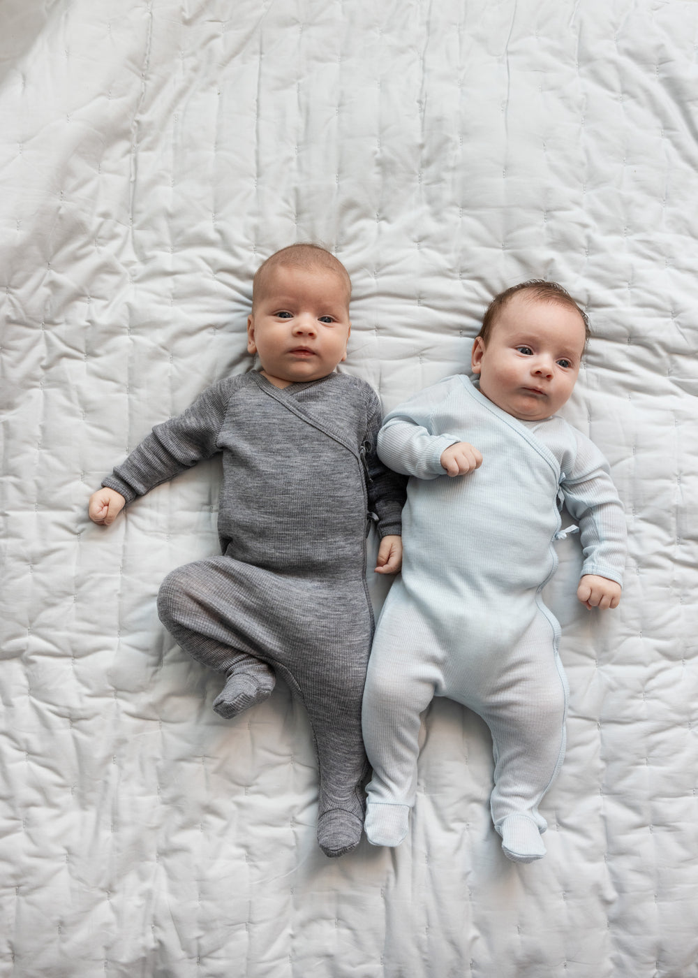 To babyer ligger på seng iført heldragter i merinould, en grå og en lyseblå