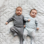 To babyer ligger på seng iført heldragter i merinould, en grå og en lyseblå
