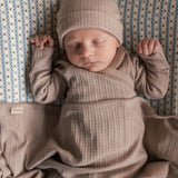 Baby iført beige pointelle sparkedragt og hue liggende med tæppe