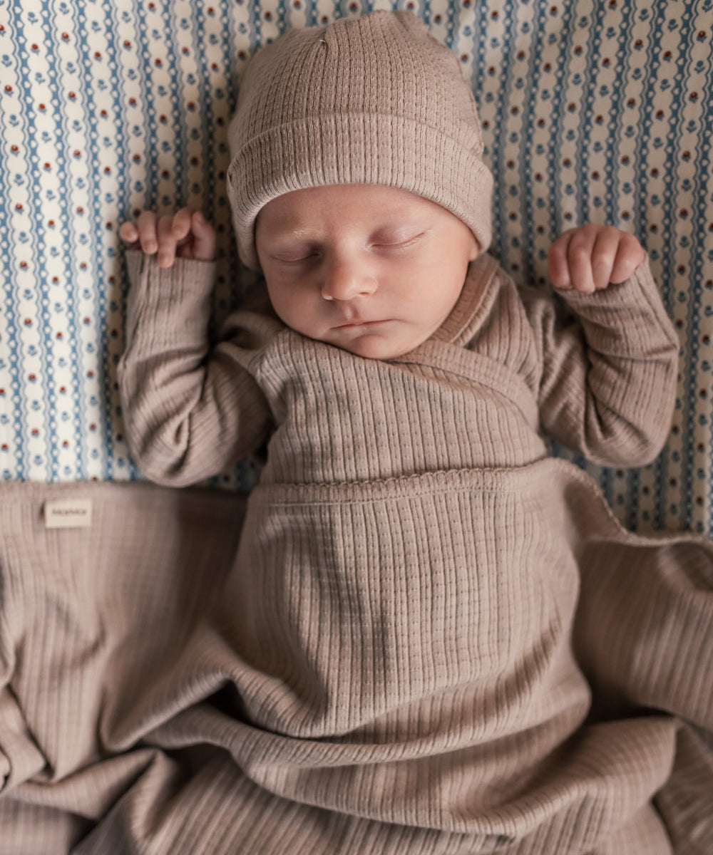 Baby iført beige pointelle sparkedragt og hue liggende på tæppe