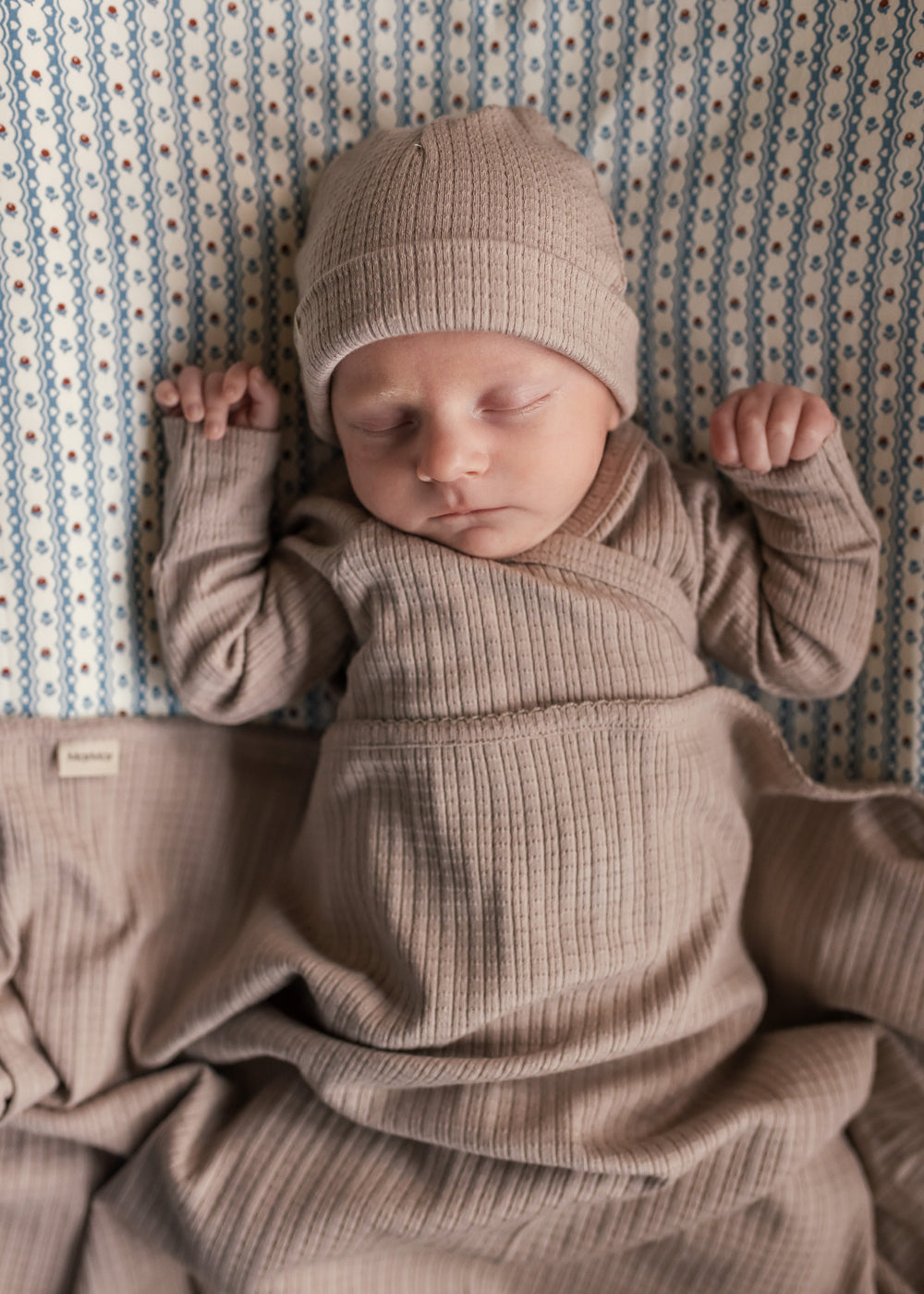 Baby iført beige pointelle sparkedragt og hue liggende på tæppe