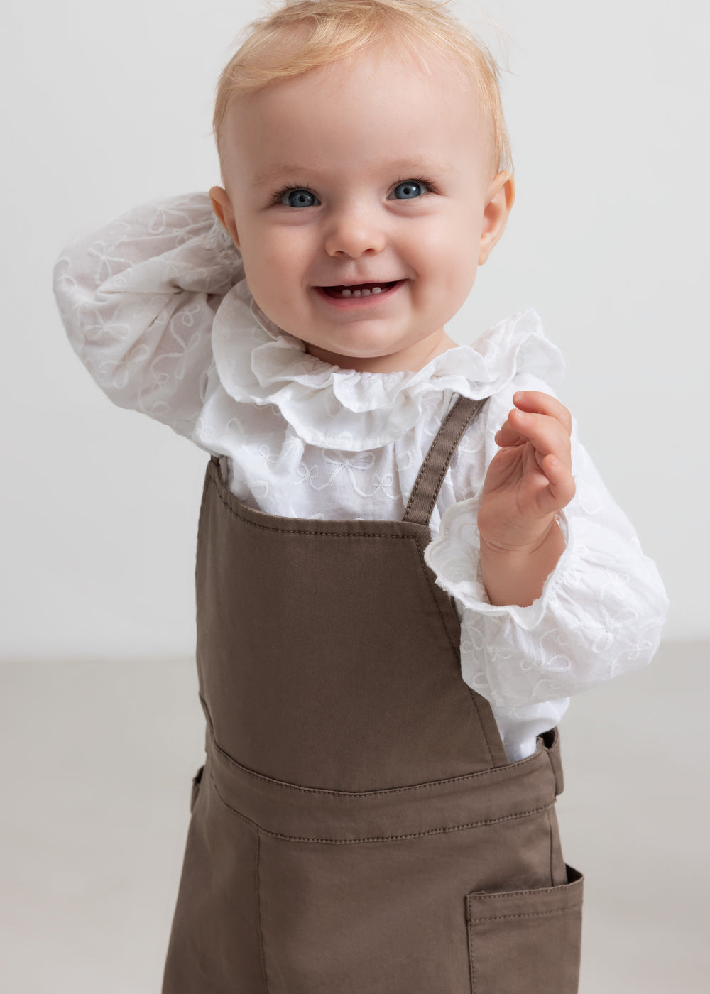 Lille børn iført hvid bluse med flæsekant og brune selebukser