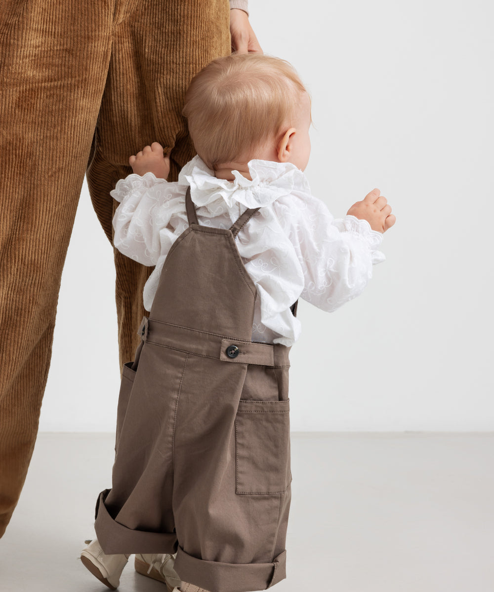 Lille børn iført hvid bluse med flæsekant og brune selebukser