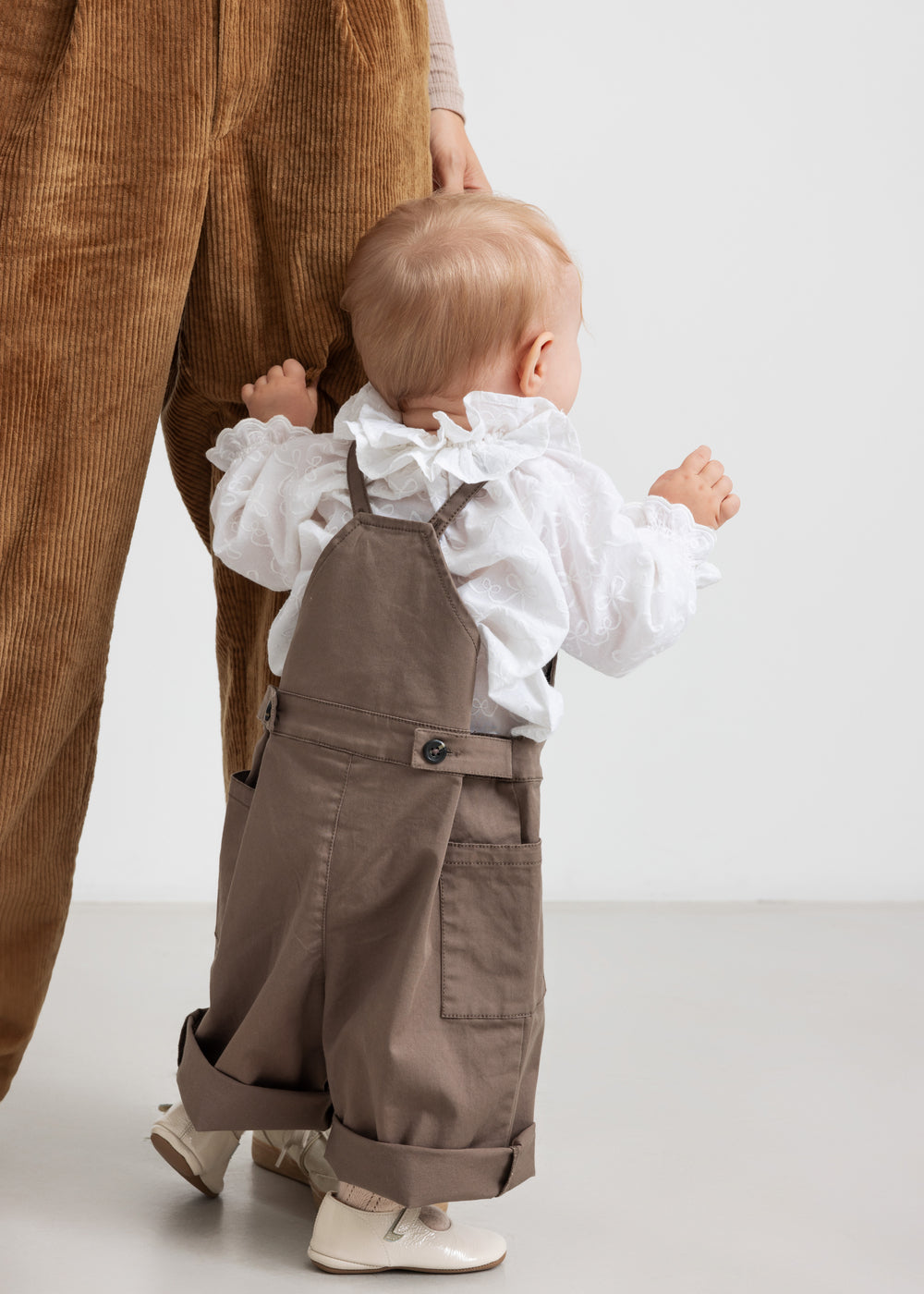 Lille børn iført hvid bluse med flæsekant og brune selebukser