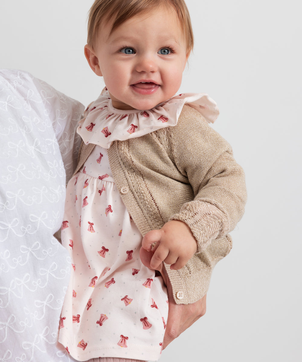 Baby iført beige cardigan med guldglimmer og lyserød julekjole med juleklokke mønster