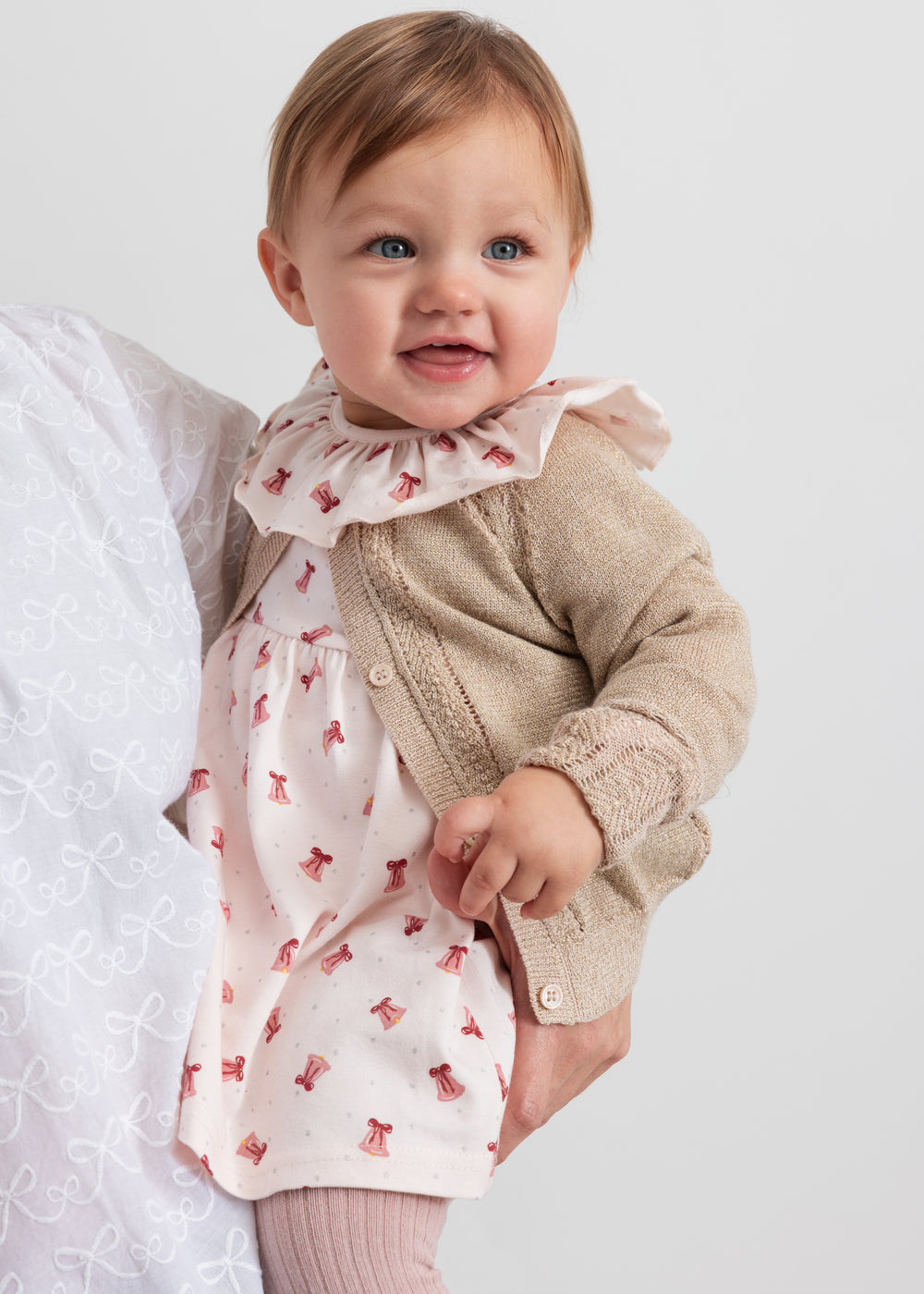Baby iført beige cardigan med guldglimmer og lyserød julekjole med juleklokke mønster