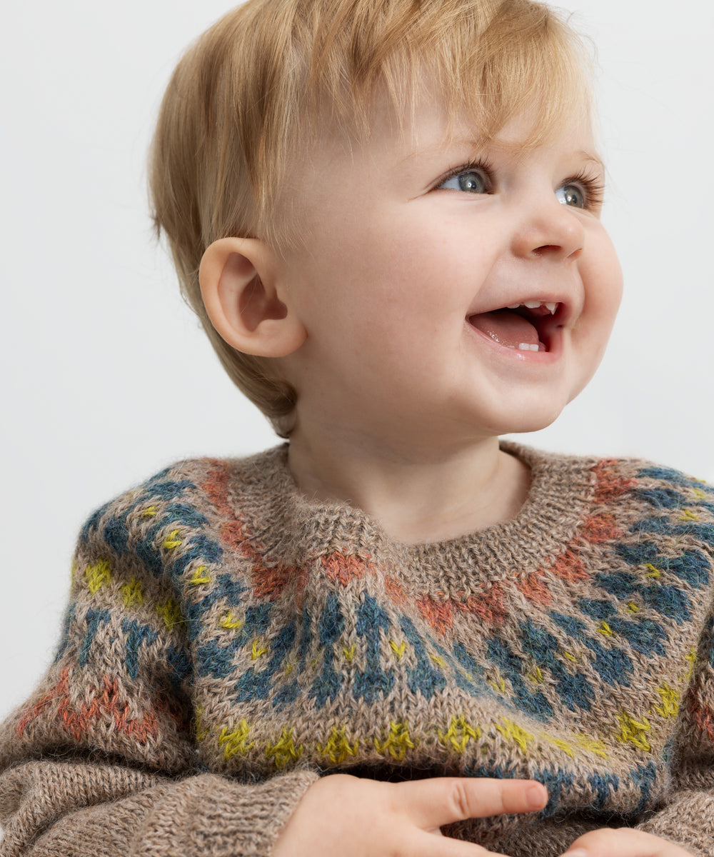 Baby smiler iført beige uldstrikbluse med farvet bærestykke