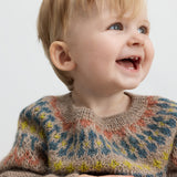 Baby smiler iført beige uldstrikbluse med farvet bærestykke