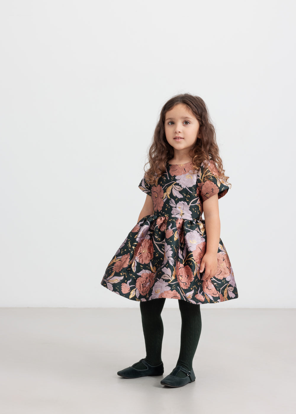 Pige iført mørkegrøn festkjole med blomsterprint og sorte strømpebukser til børn