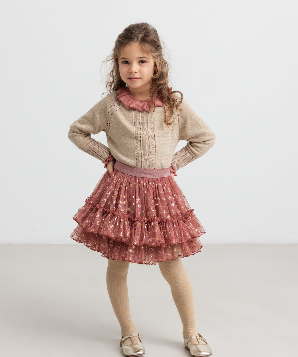 Pige iført beige cardigan med guldglimmer, lyserød festnederdel og strømpebukser til børn