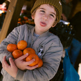 Barn iført brun strikket hue og jakke med frugt i hænderne