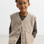 Barn iført lys teddyvest med hænderne i lommerne og brun t-shirt under