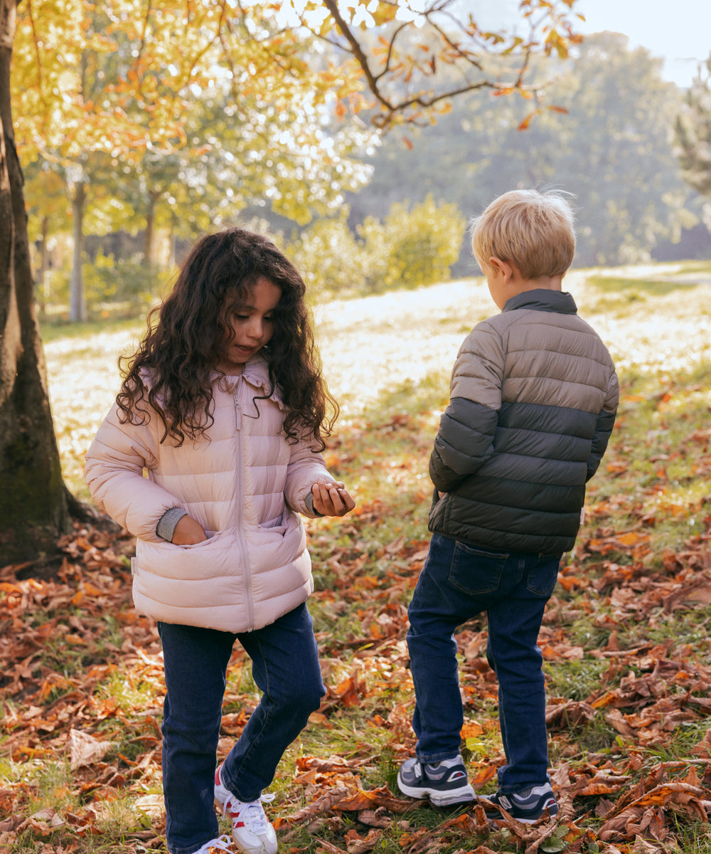 To børn leger i efterårsblade iført vatterede jakker, en lyserød og en grøn