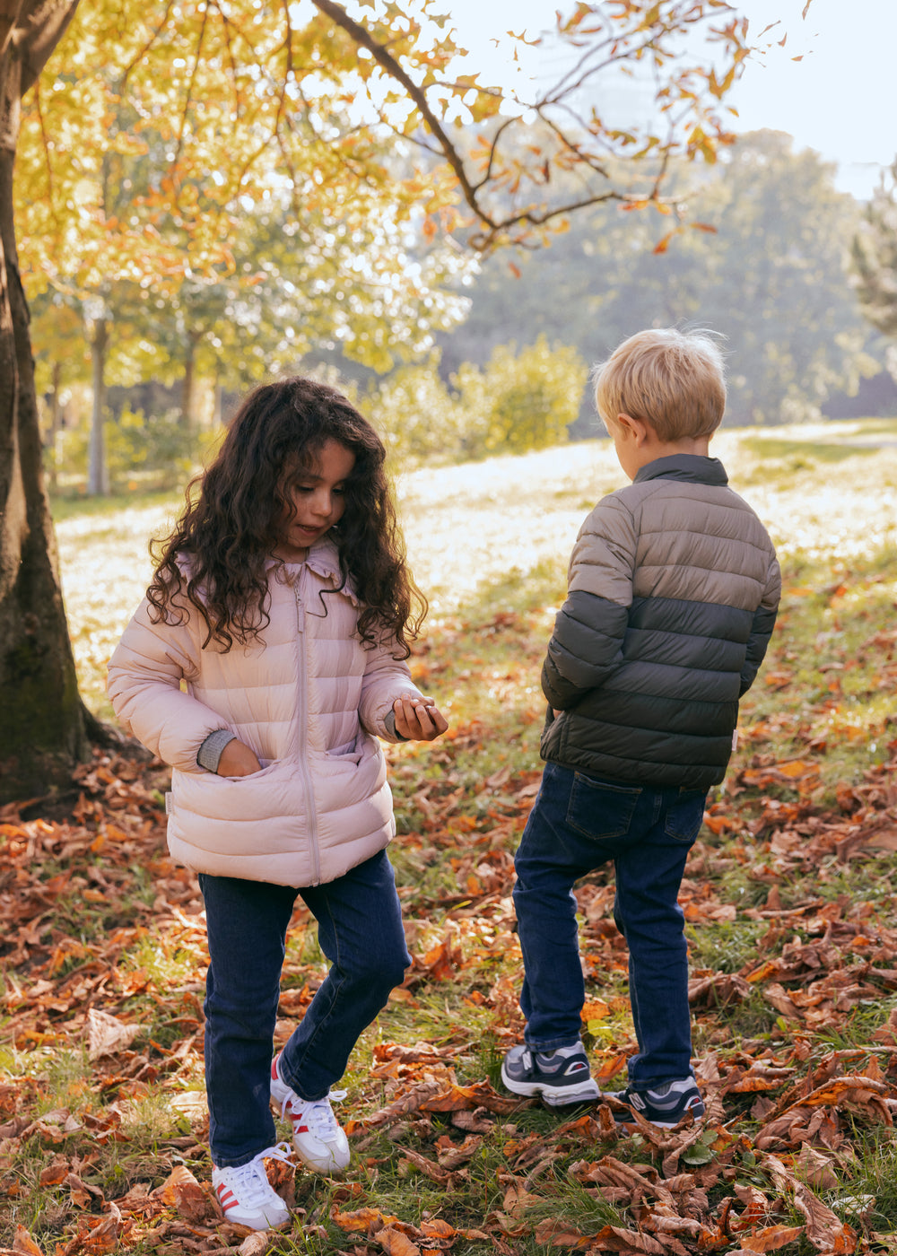 To børn leger i efterårsblade iført vatterede jakker, en lyserød og en grøn