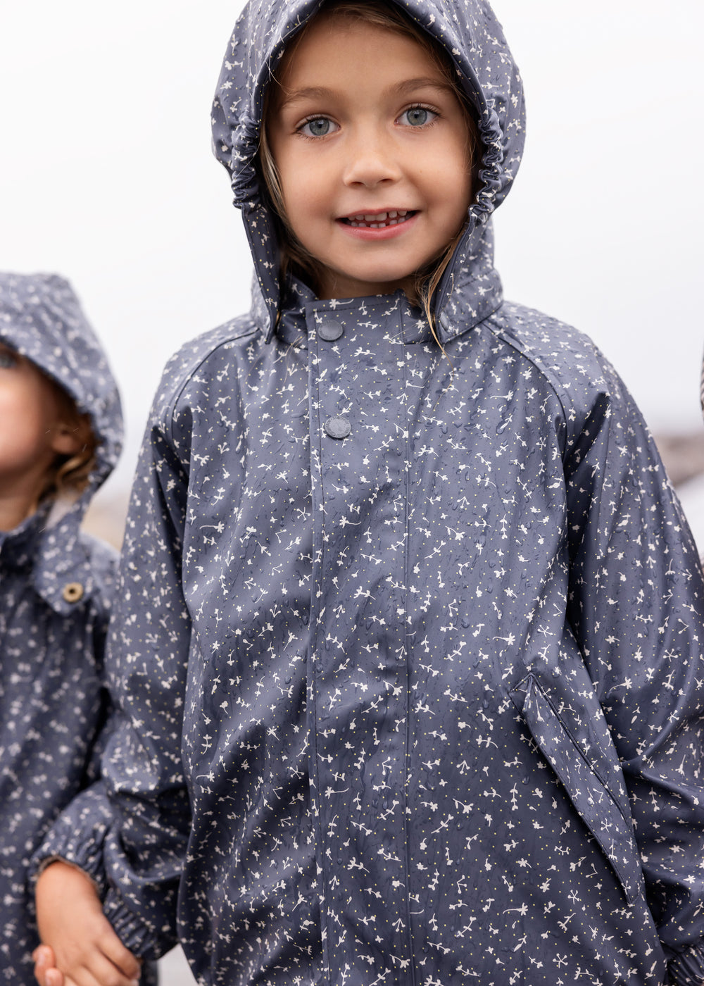 Barn iført mørkeblåt regnsæt med hætte, smilende og kigger op