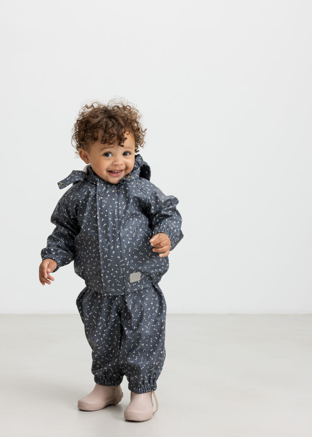 Baby i mørkeblåt regnsæt med småprint og hætte, smilende og iført lyserøde gummistøvler