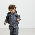 Baby i mørkeblåt regnsæt med småprint og hætte, smilende og iført lyserøde gummistøvler