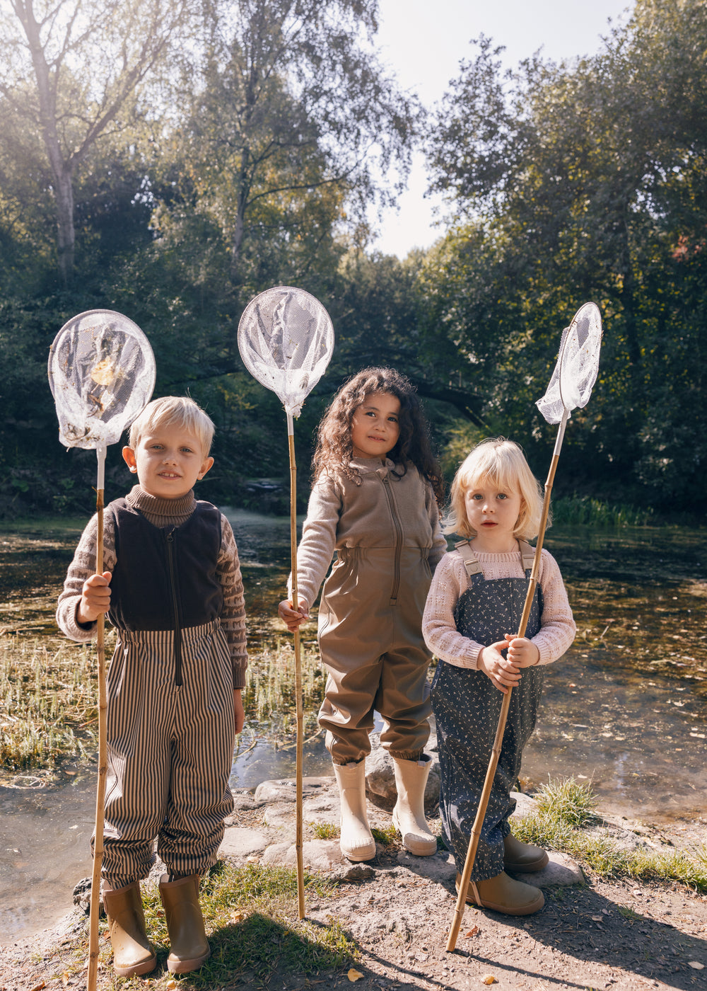 Tre børn i naturen iført regntøj, to med prikmønster og fiskenet