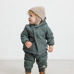 Baby iført støvet grøn termojakke og bukser, står og kigger til siden