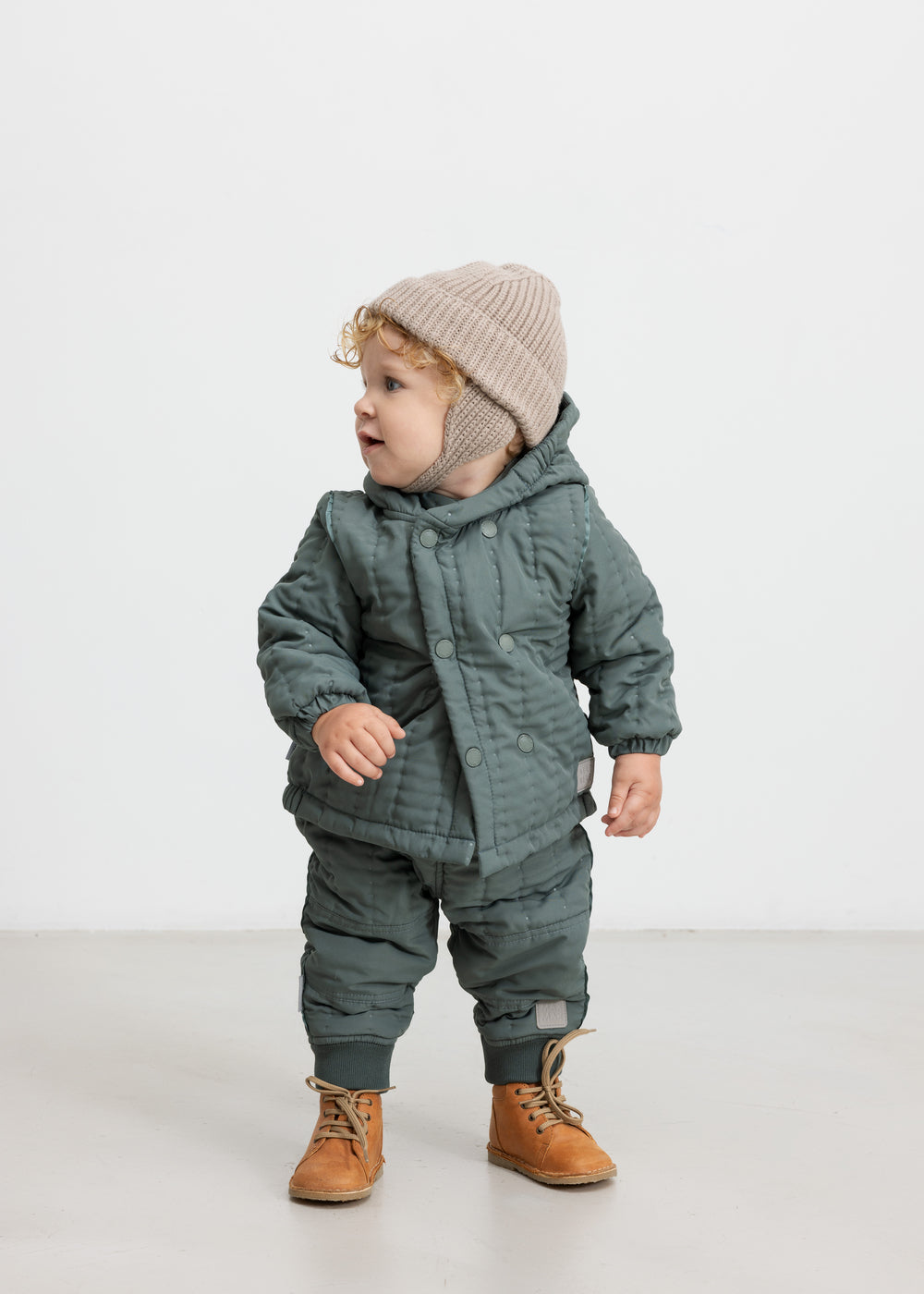 Baby iført støvet grønt termotøj og beige hue, står og kigger til siden