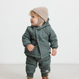 Baby iført støvet grønt termotøj og beige hue, står og kigger til siden