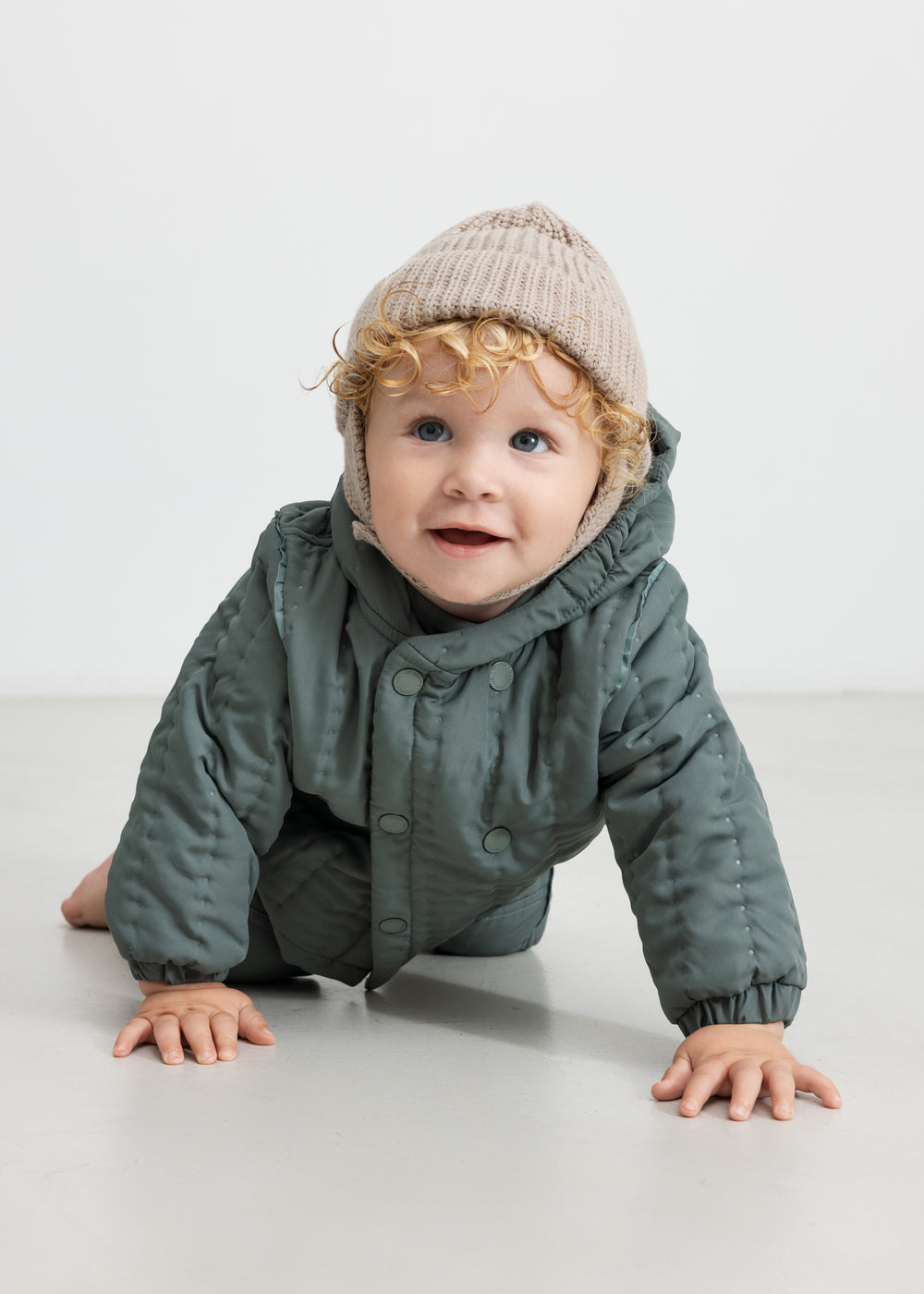 Baby iført beige strikhue og mørkegrøn heldragt, kravler mod kameraet