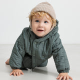 Baby iført beige strikhue og mørkegrøn heldragt, kravler mod kameraet