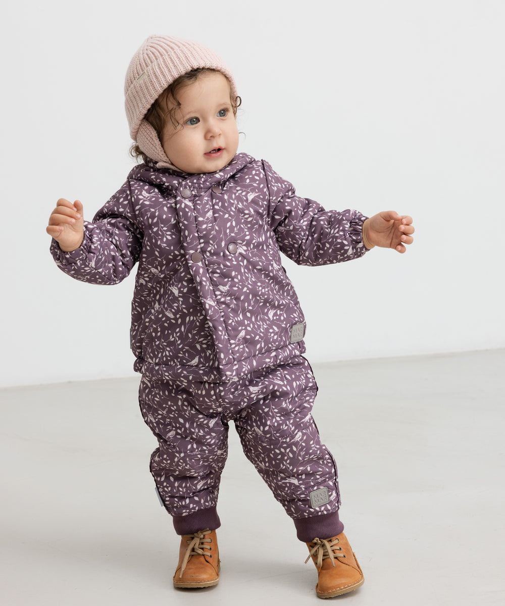 Baby iført lyserød strikhue og lilla heldragt, står og smiler