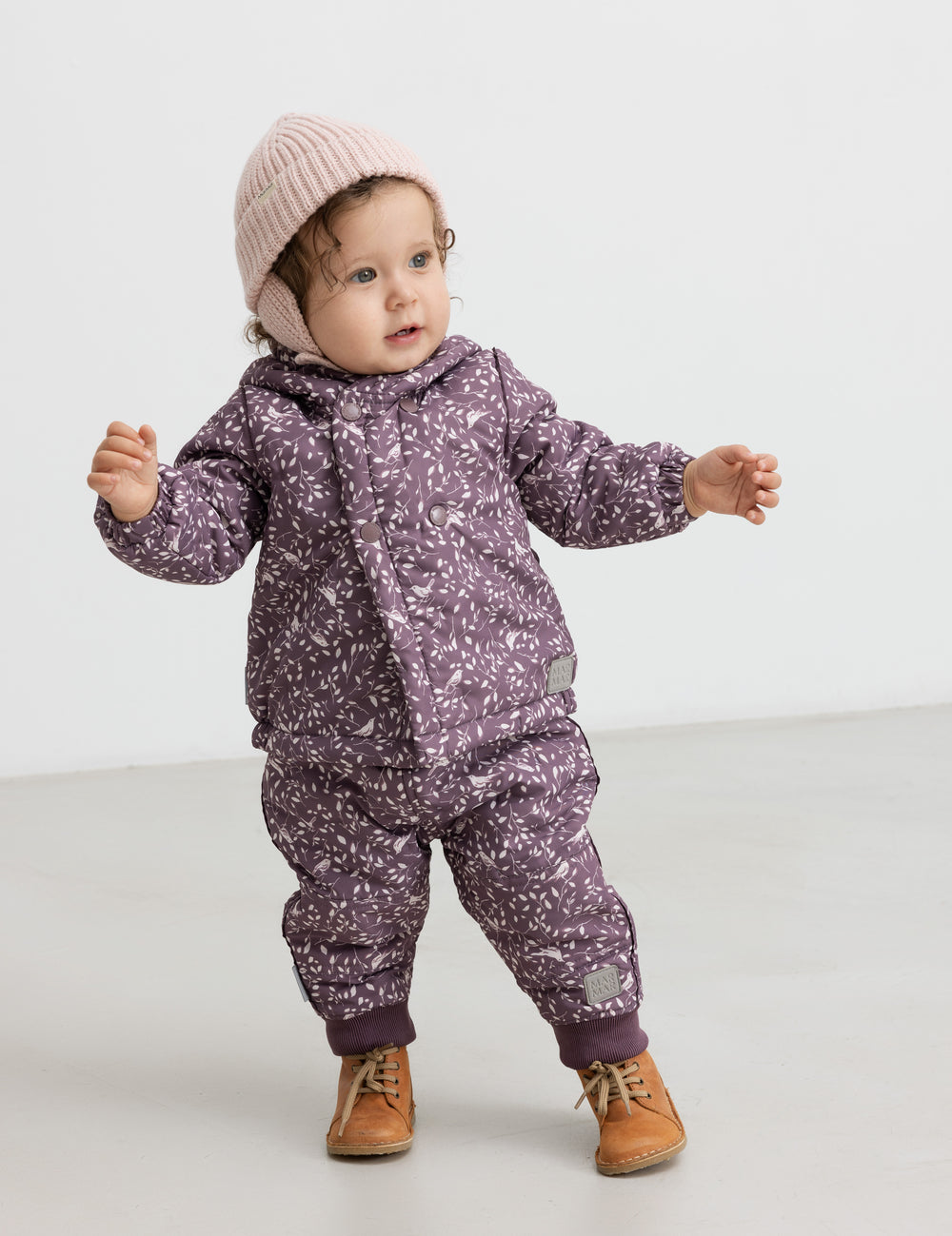 Baby iført lyserød strikhue og lilla heldragt, står og smiler.