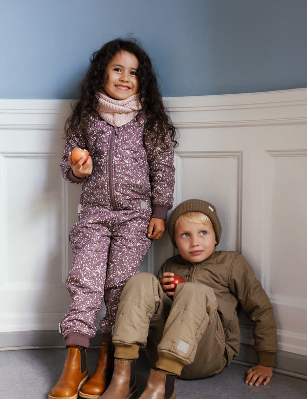 To børn iført termotøj og boots sidder og står indendørs med æbler