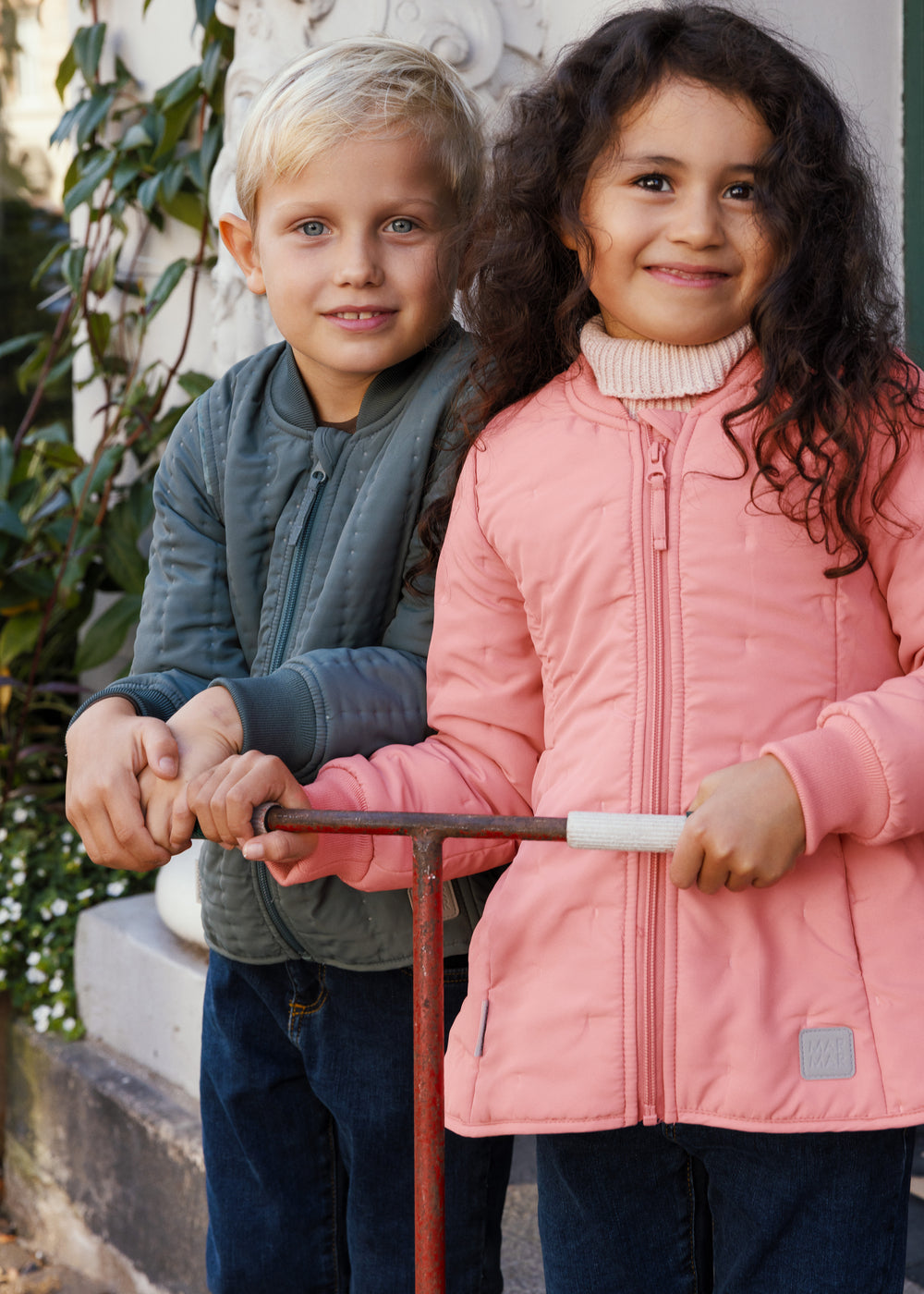 Pige i lyserød termojakke står med scooter ved siden af en dreng