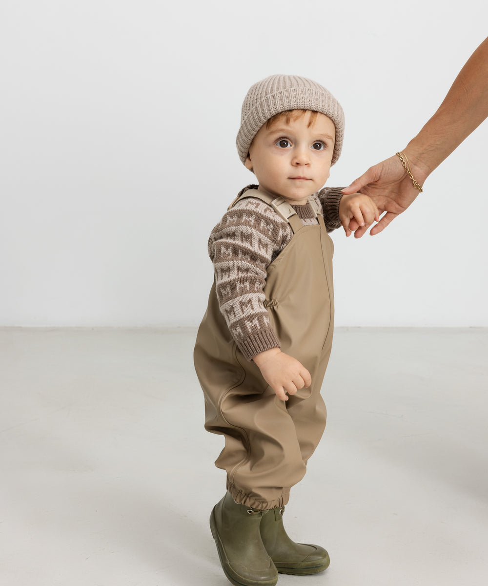 Baby iført brun M-mønstret striktrøje, termobukser og hue, holder en voksen i hånden
