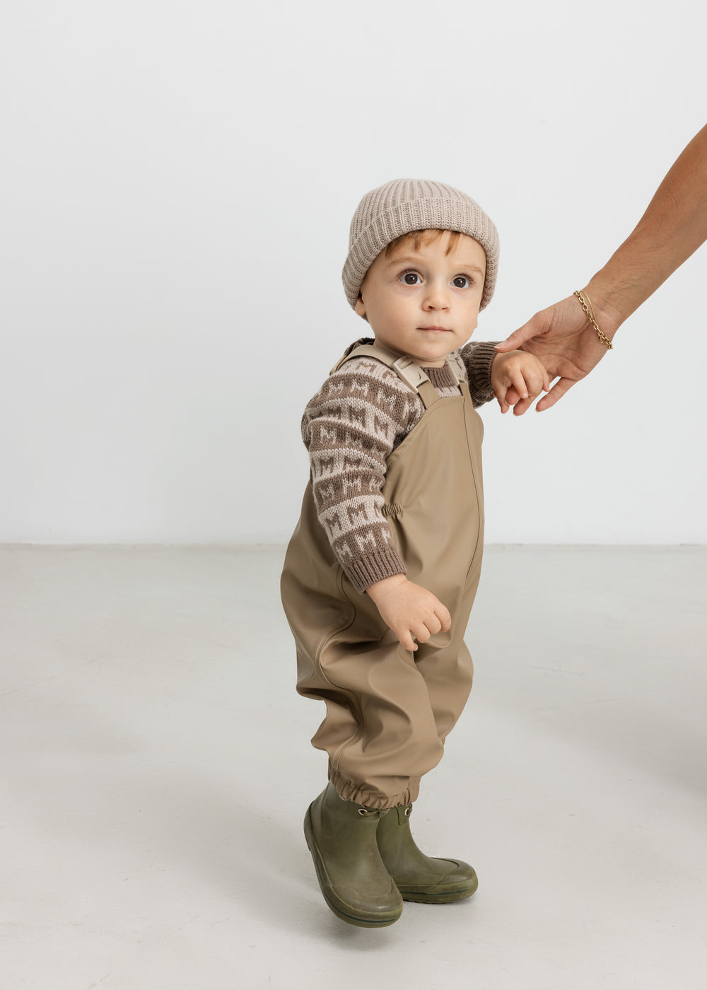 Baby iført brun M-mønstret striktrøje, termobukser og hue, holder en voksen i hånden