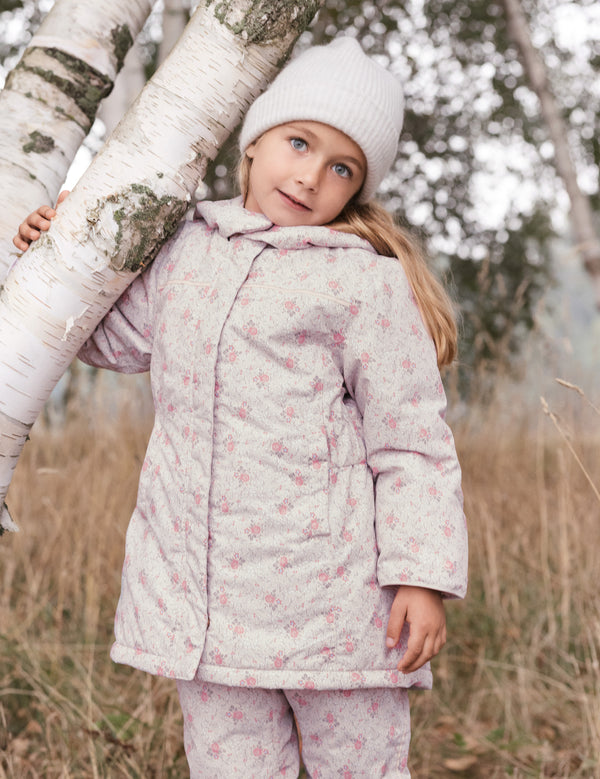 Pige iført lyserødt blomstret vinterovertøj og hvid hue ved et birketræ