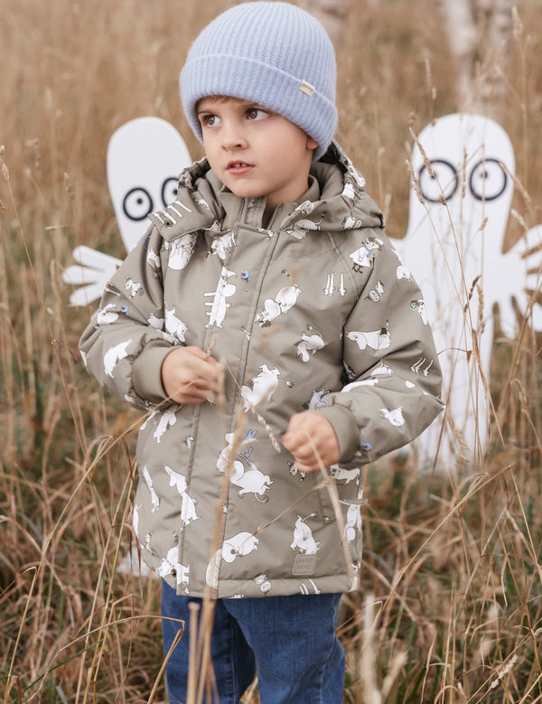 Dreng iført Moomin mønstret vinterjakke og lyseblå hue står i højt græs