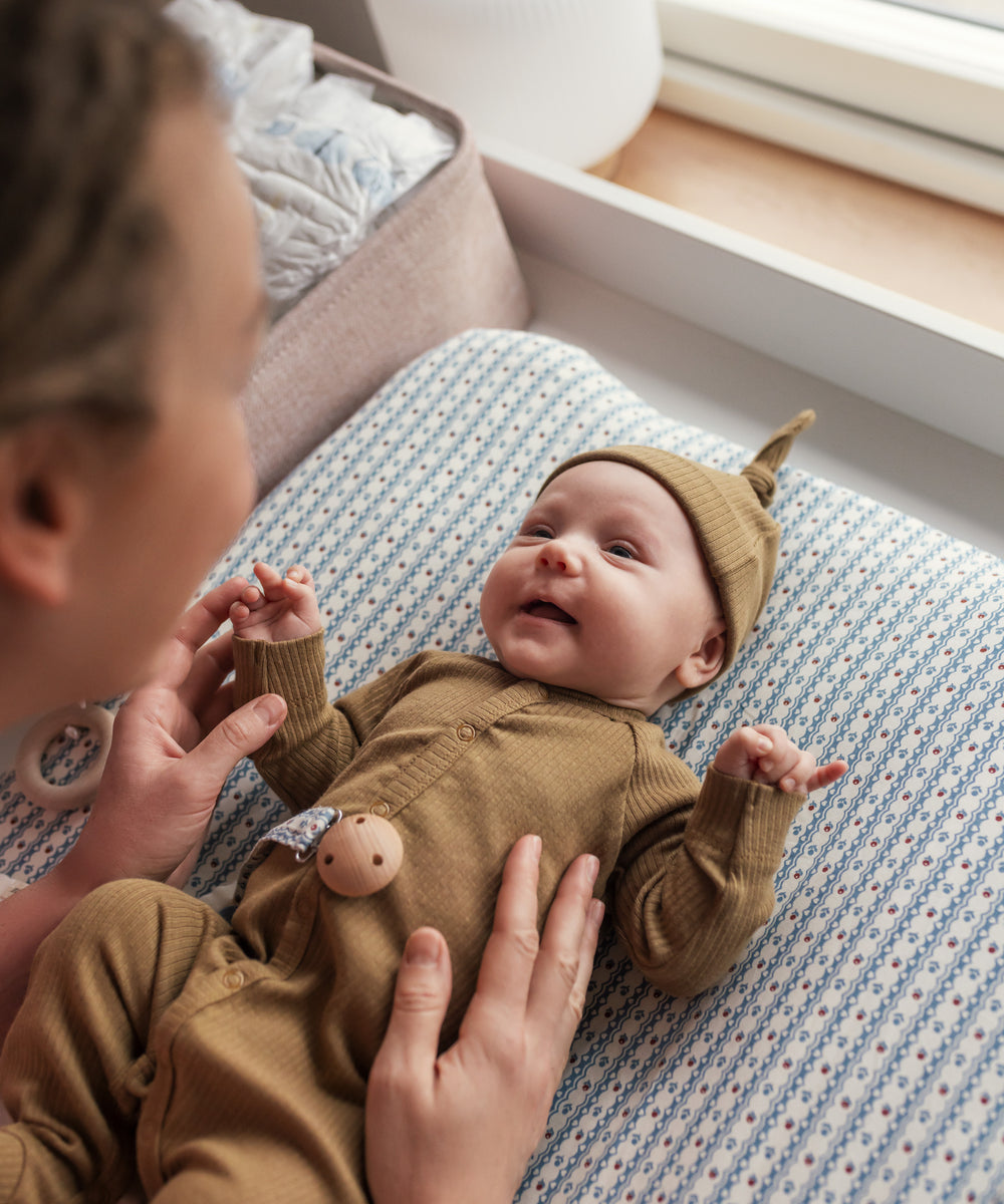 Mor leger med baby iført brun ribstrikket hue og heldragt ved puslebord.