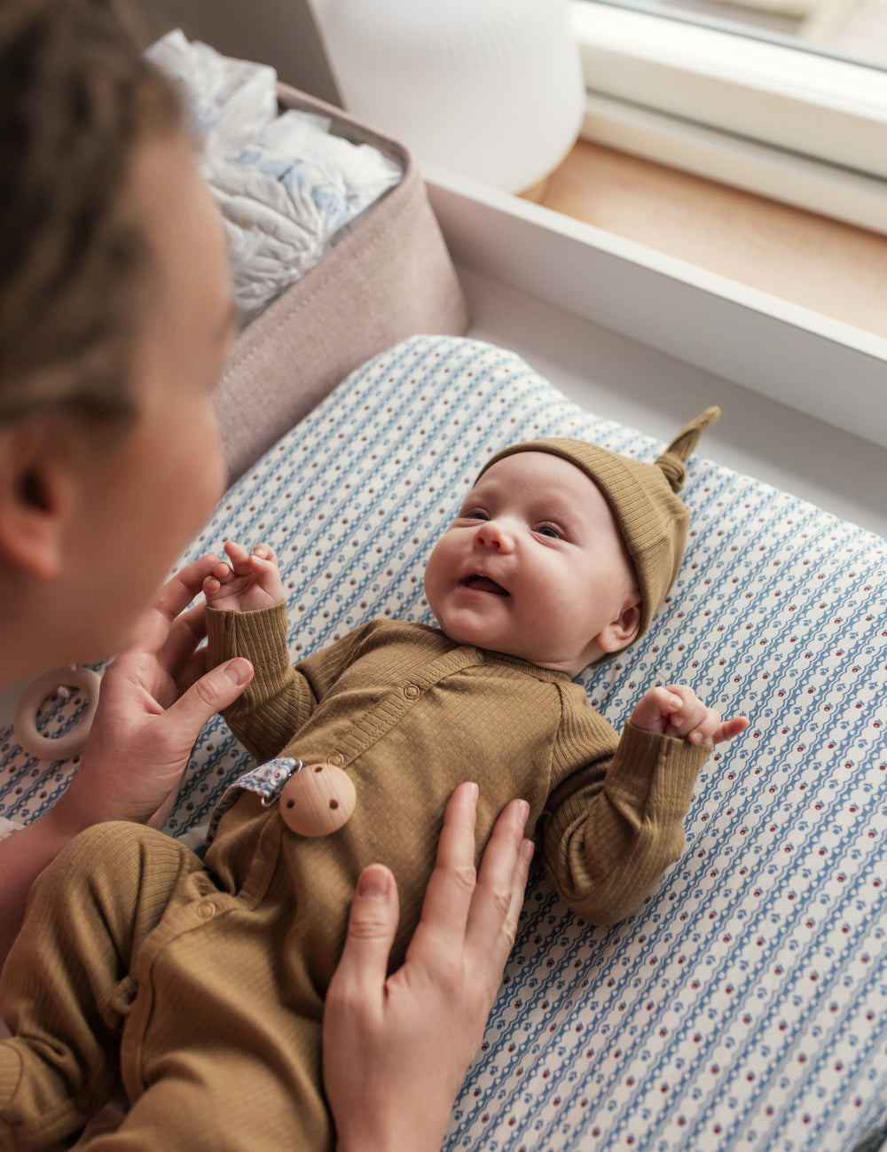 Mor leger med baby iført brun ribstrikket hue og holdt ved puslebord.