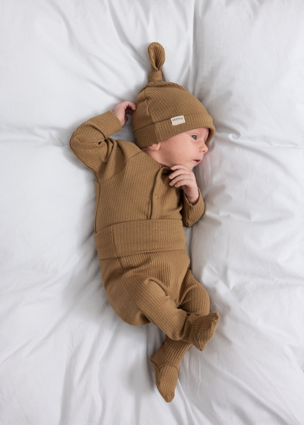 Baby med brun ribstrikket hue og matchende heldragt ligger på hvidt lagen