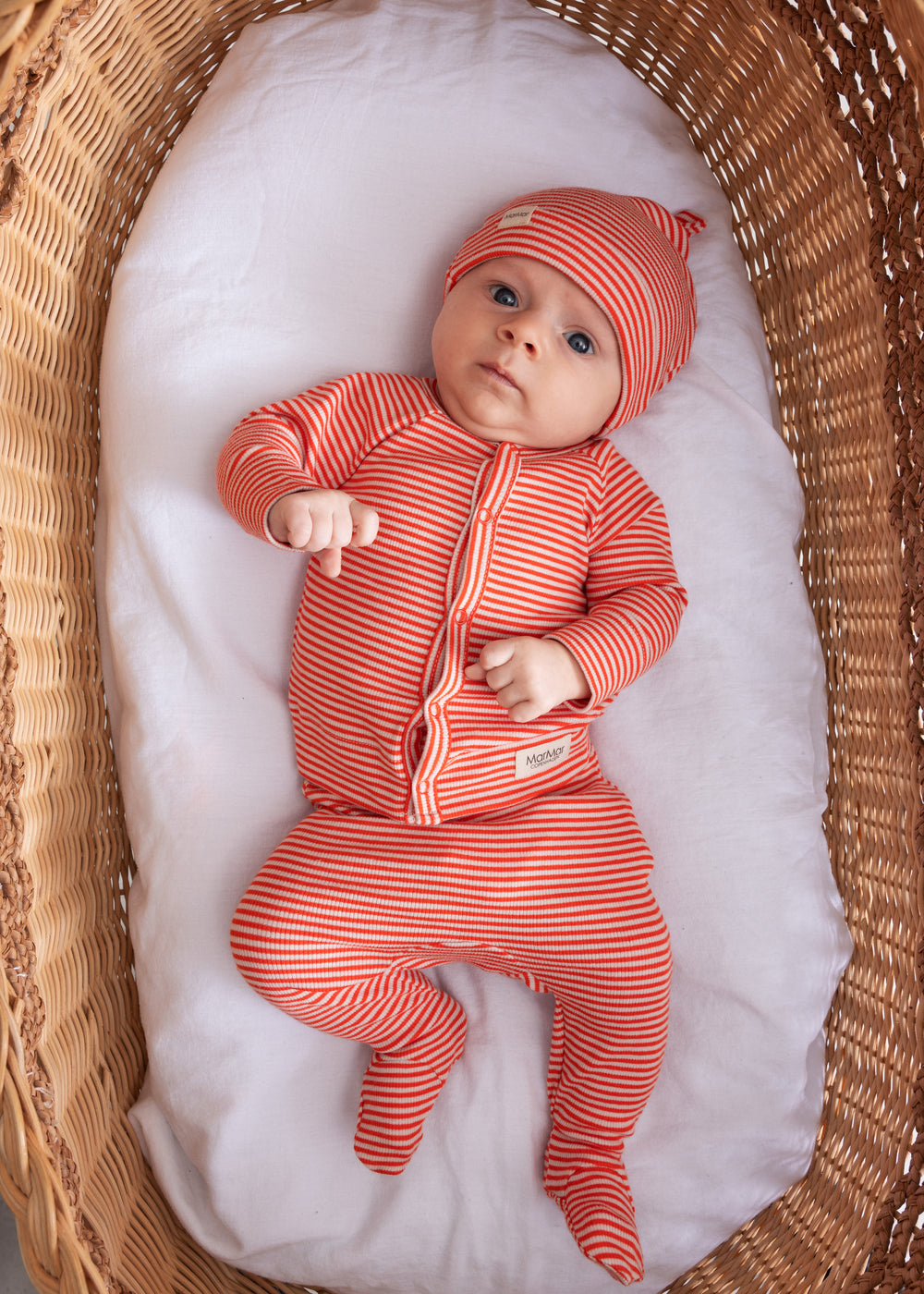 Baby iført koralfarvet stribet sæt med hue, cardigan og bukser i en fletkurv