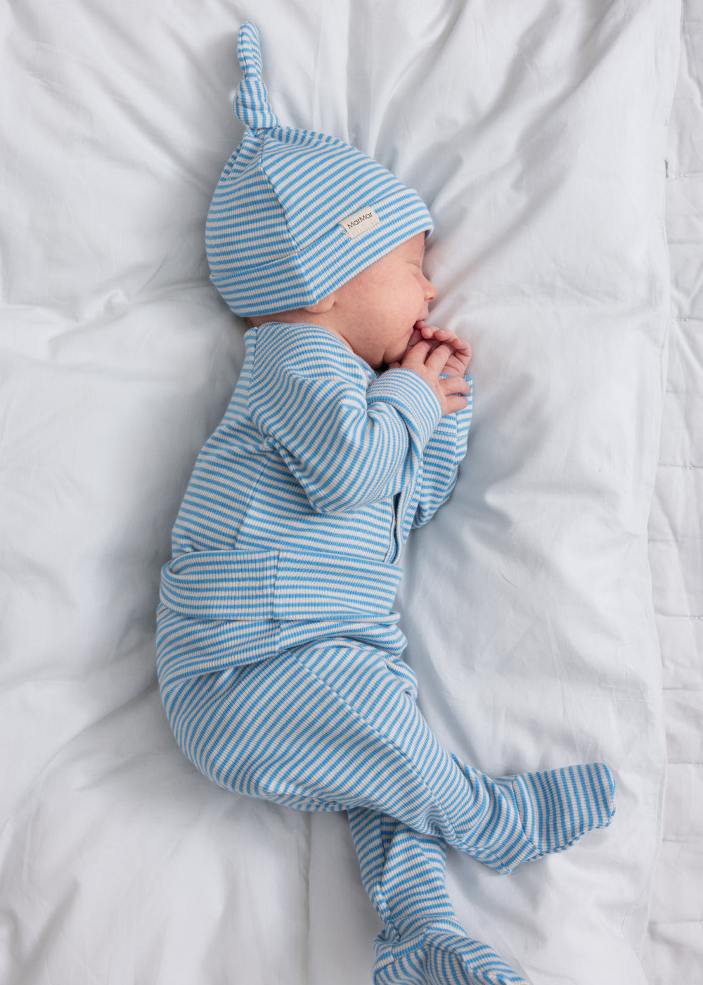 Baby iført blå- og hvidstribet cardigan og hue, liggende på hvidt tæppe